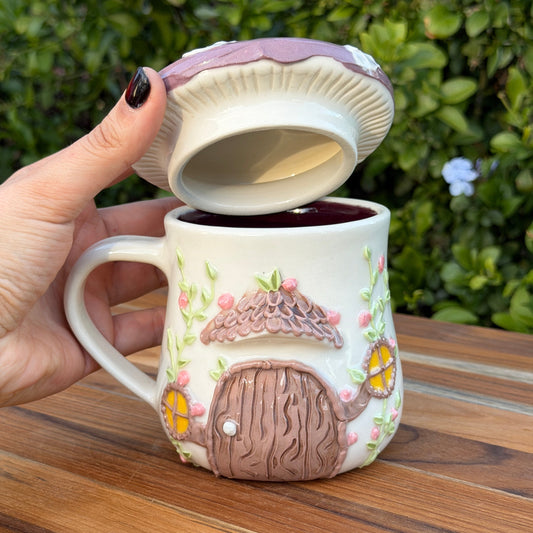 Mushroom-shaped mug with a lid, decorated with a house design, held by a hand on a wooden surface with greenery in the background.